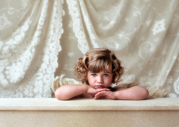 Studio portrait of a little girl, arms on table, hands under chin, with wings on her back, cherub-like.